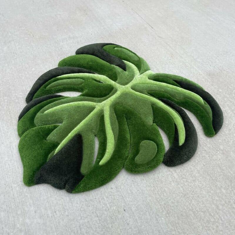 Green and black monstera leaf-shaped rug on light gray surface.