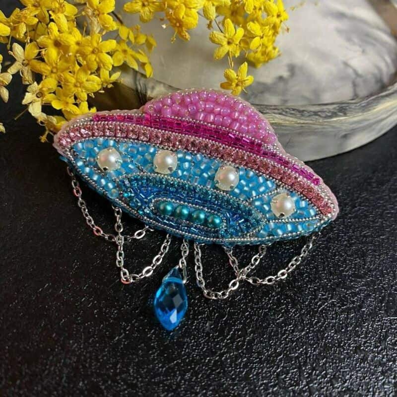 Pink and blue beaded eye-shaped brooch with dangling chains and a blue teardrop pendant, placed on a.
