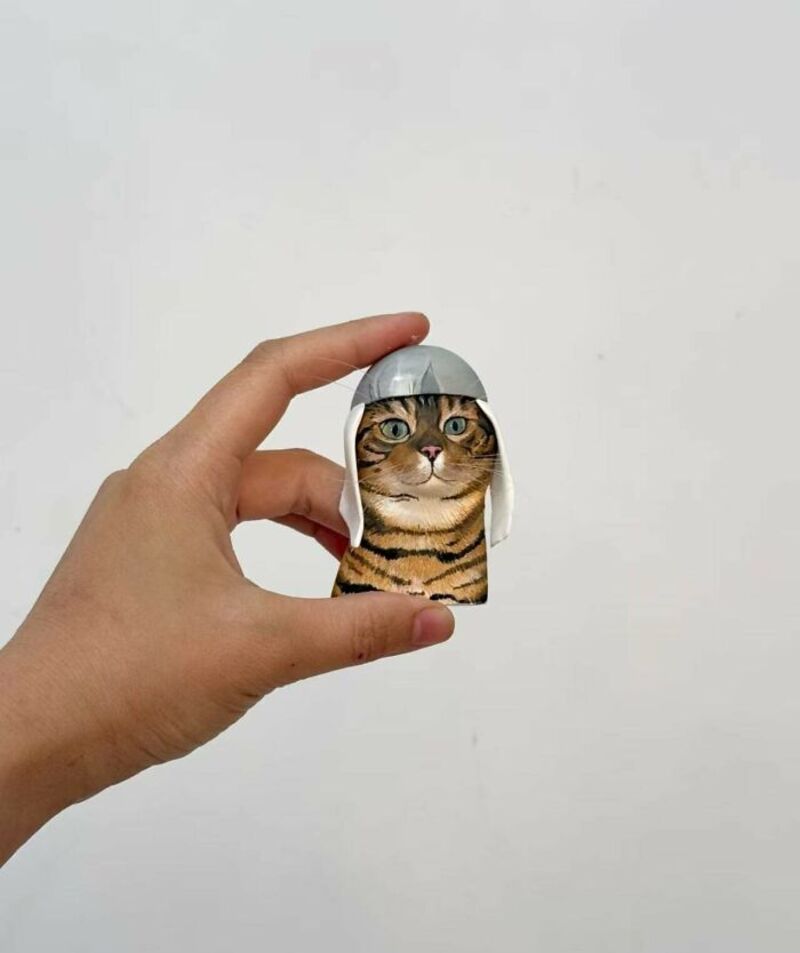 Cat puppet with a gray helmet held by hand against a plain white background.