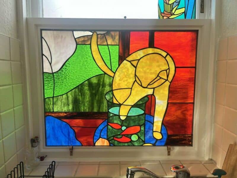Stained glass window with a yellow cat and a fish bowl, colorful background.