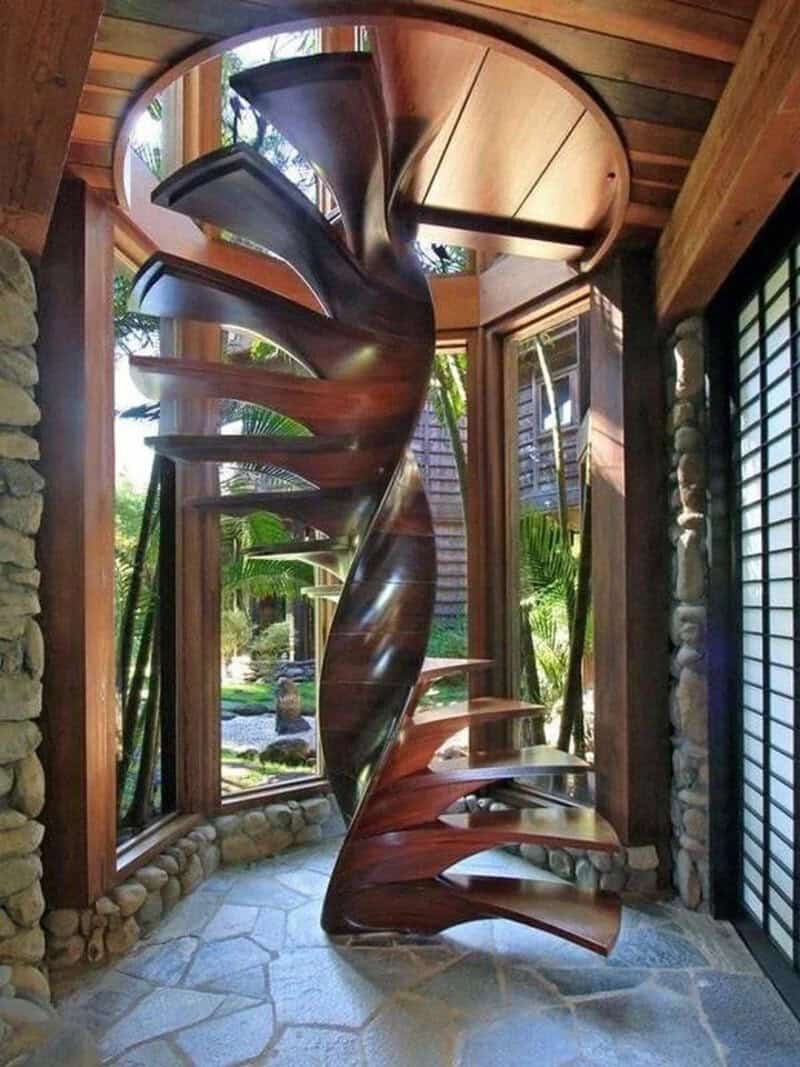 Spiral wooden staircase inside a house with large windows and stone walls.