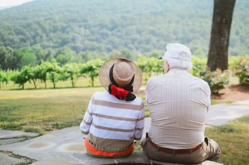 Older man and woman sitting on a bench in a park, facing away from the camera.