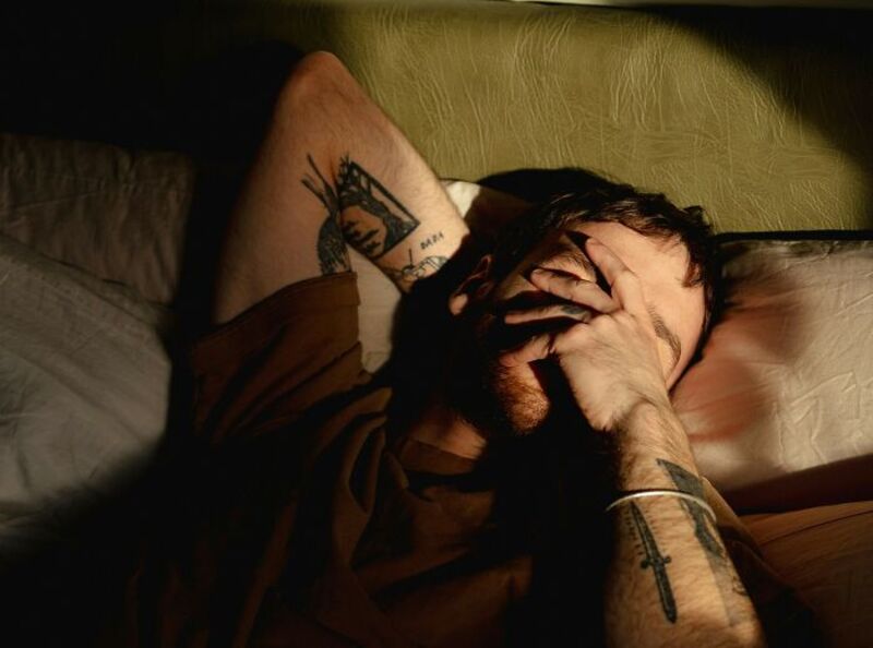 Man with tattoos covering his face and arms, lying on a bed with his hand over his face.