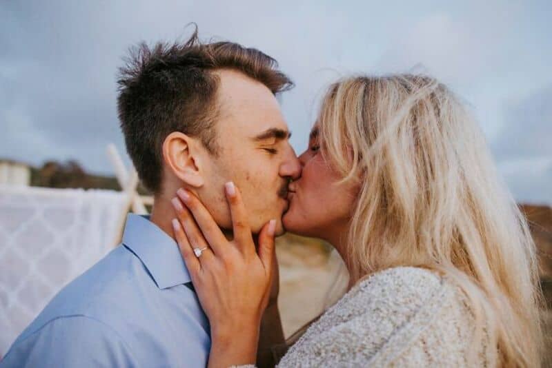 A couple kissing outdoors, with a cloudy sky in the background.