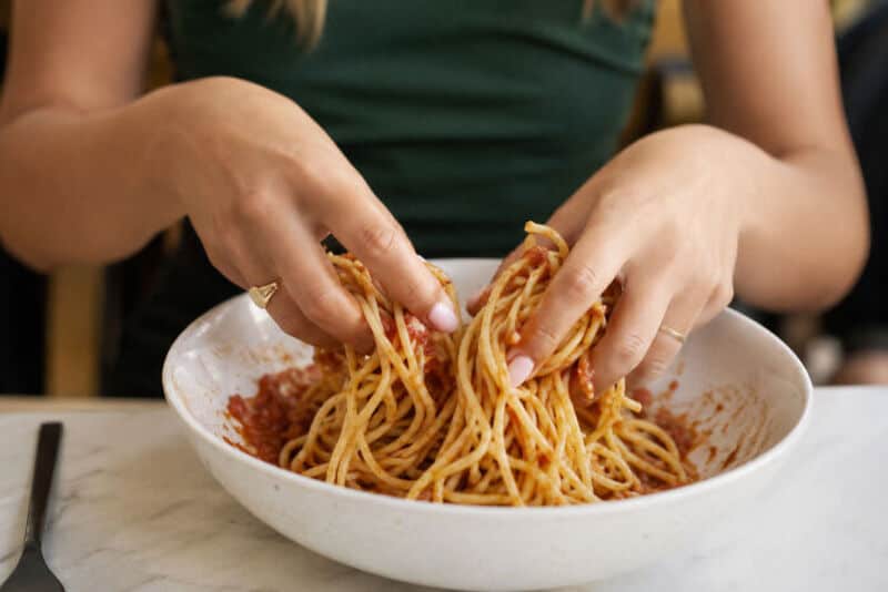 Person mixing spaghetti with hands in a bowl of sauce.