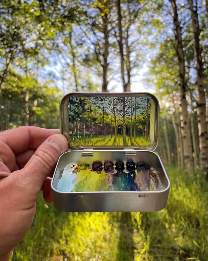 Hand holding a small metal case with a painted forest scene inside, outdoors in a wooded area.