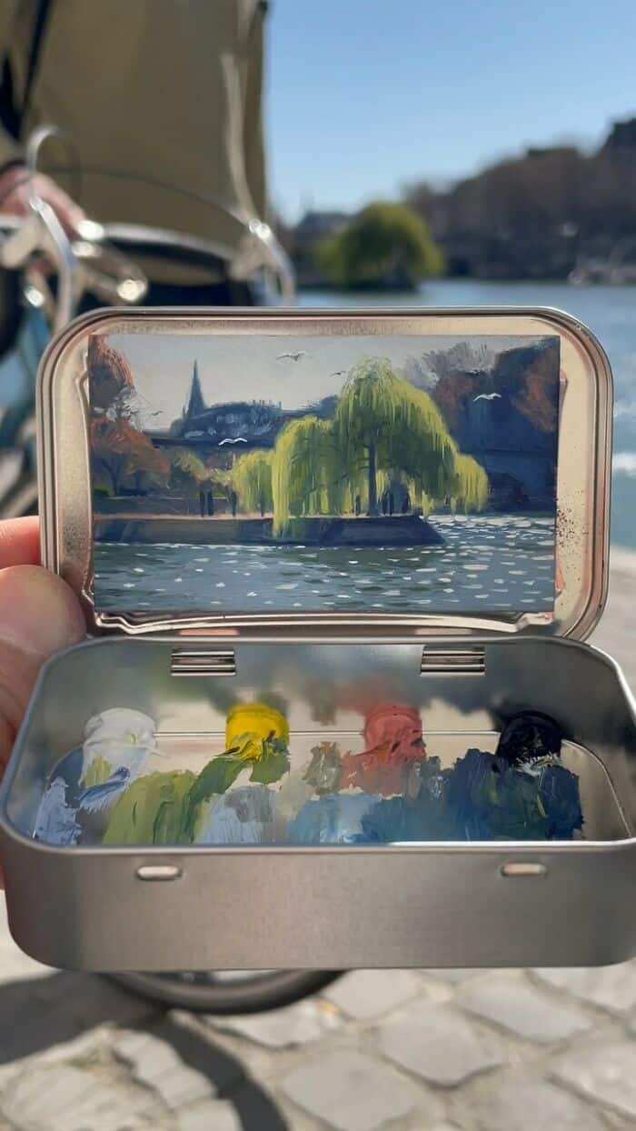 Painting of a riverside scene with a church and a weeping willow tree in a portable case.