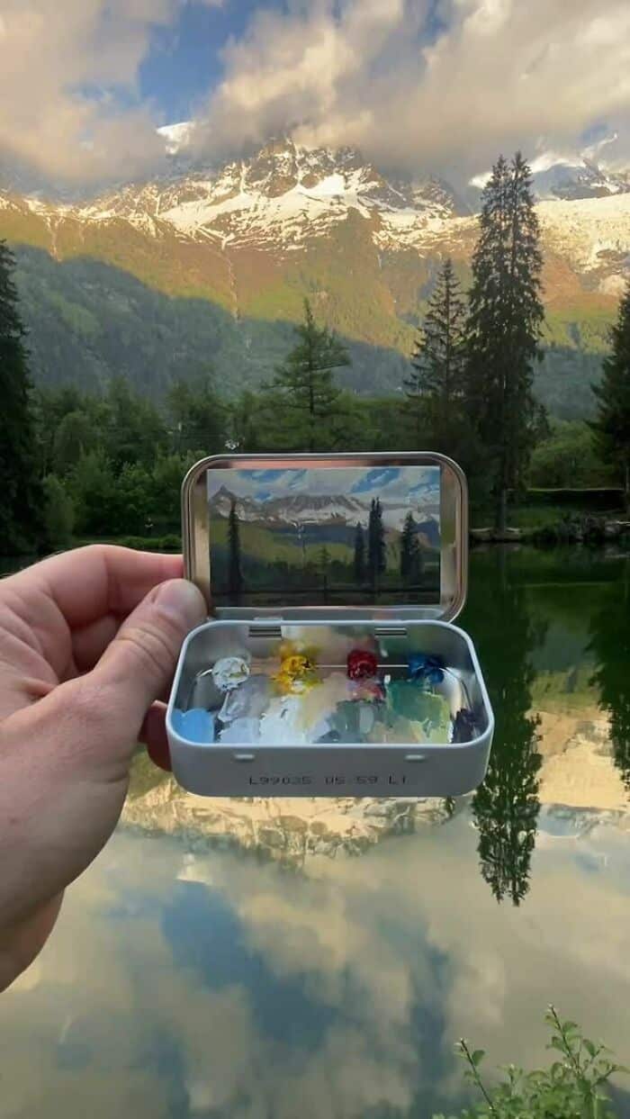 A small metal case with a mirror reflecting a mountain landscape and a hand holding it.
