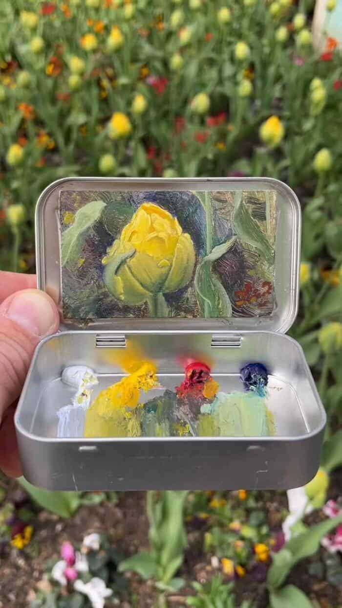 Yellow flower inside a small, open metal container with a mirror lid, set against a background of co.