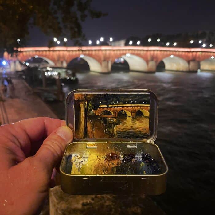 Hand holding a small metal box with a mirror, reflecting a bridge over a river at night.