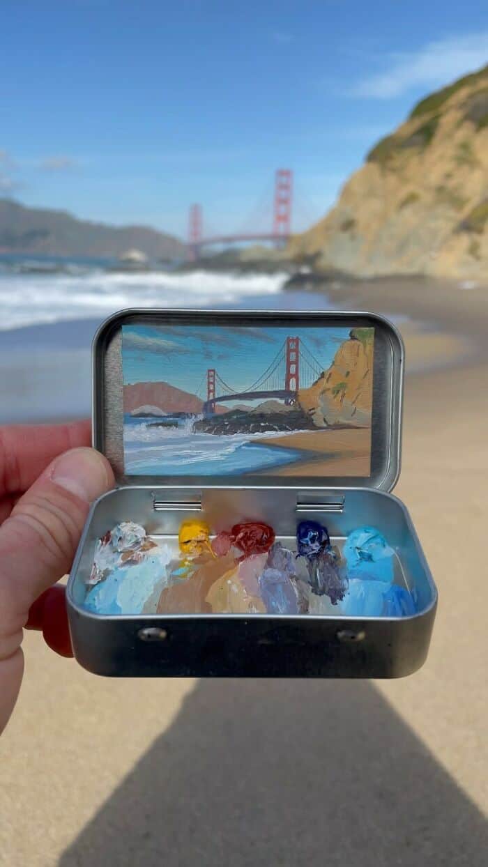 Hand holding a case with a painting of the Golden Gate Bridge on a beach.