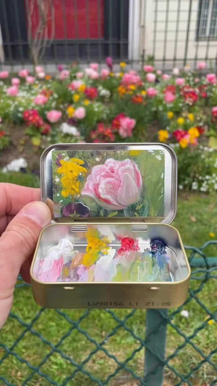 Pink tulip in a small metal box with colorful flowers painted inside, held in front of a garden with.