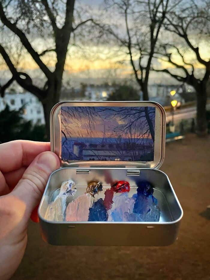Hand holding a metal box with paint and a mirror, outdoors at sunset with trees and cityscape.