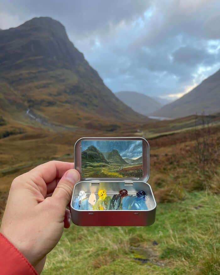Hand holding a small box with colorful glass art pieces, mountain landscape background.