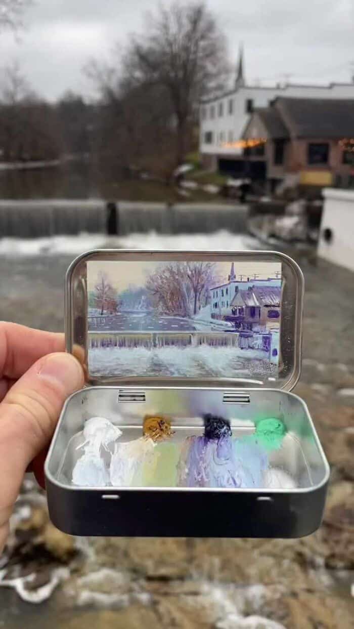 Hand holding a small case with colorful paint blobs, river and buildings in background.
