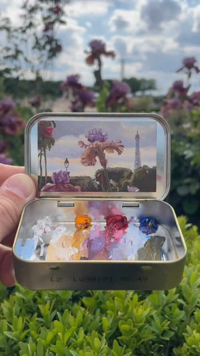 A colorful palette of paint inside a small container with a scenic background of flowers and a tower.