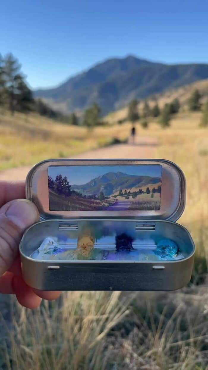 Hand holding a small container with a mirror reflecting a mountain landscape.