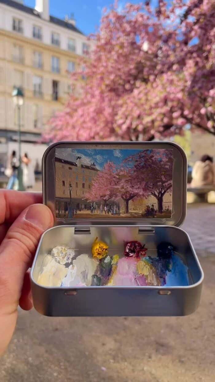 Hand holding a small box with colorful flowers and a pink tree reflected in a phone screen.