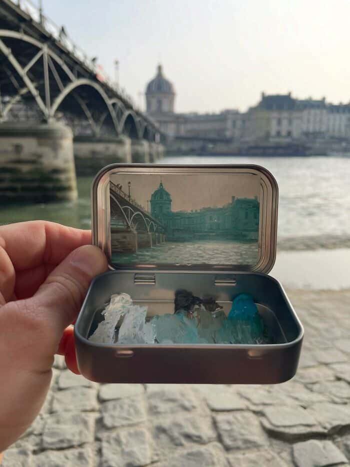 Hand holding a small container with water and rocks, with a cityscape and bridge in the background.
