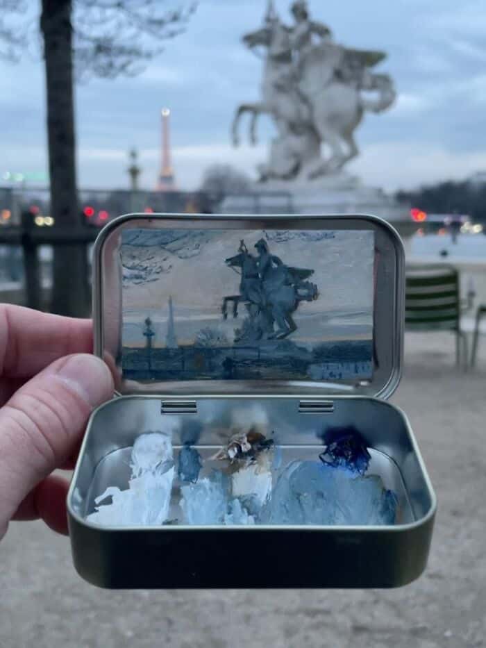 Miniature snow scene with a tiny figure and trees inside a metal box, with a large statue in the bac.