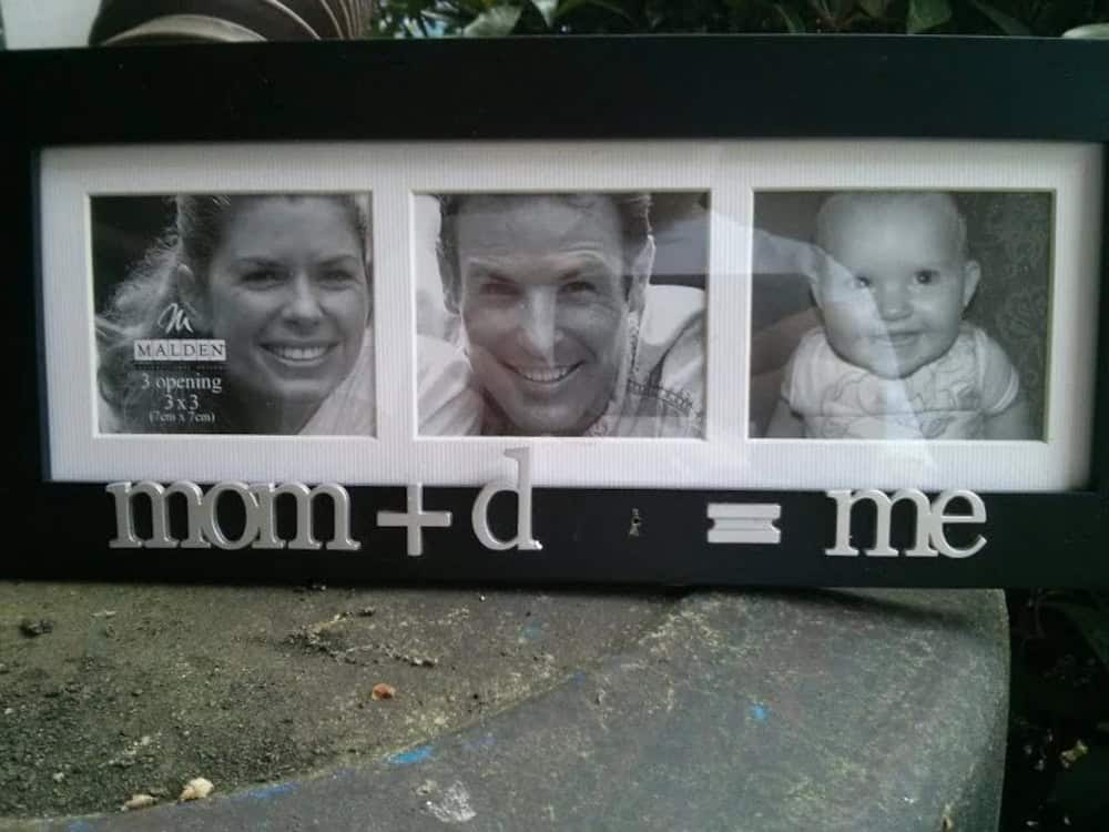 Family photo with mom, dad, and baby in a black frame with "mom + d = me" text.