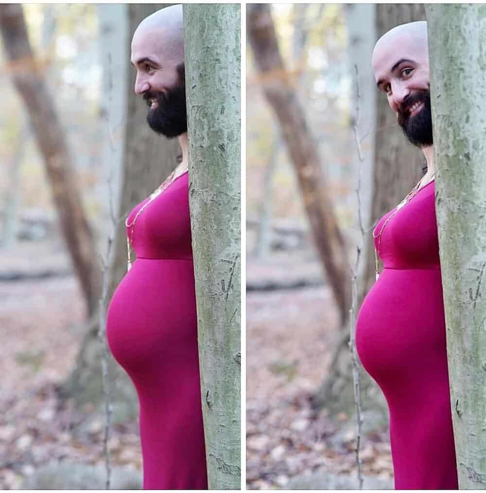 Woman with a pregnant belly standing behind a tree outdoors in a pink dress.
