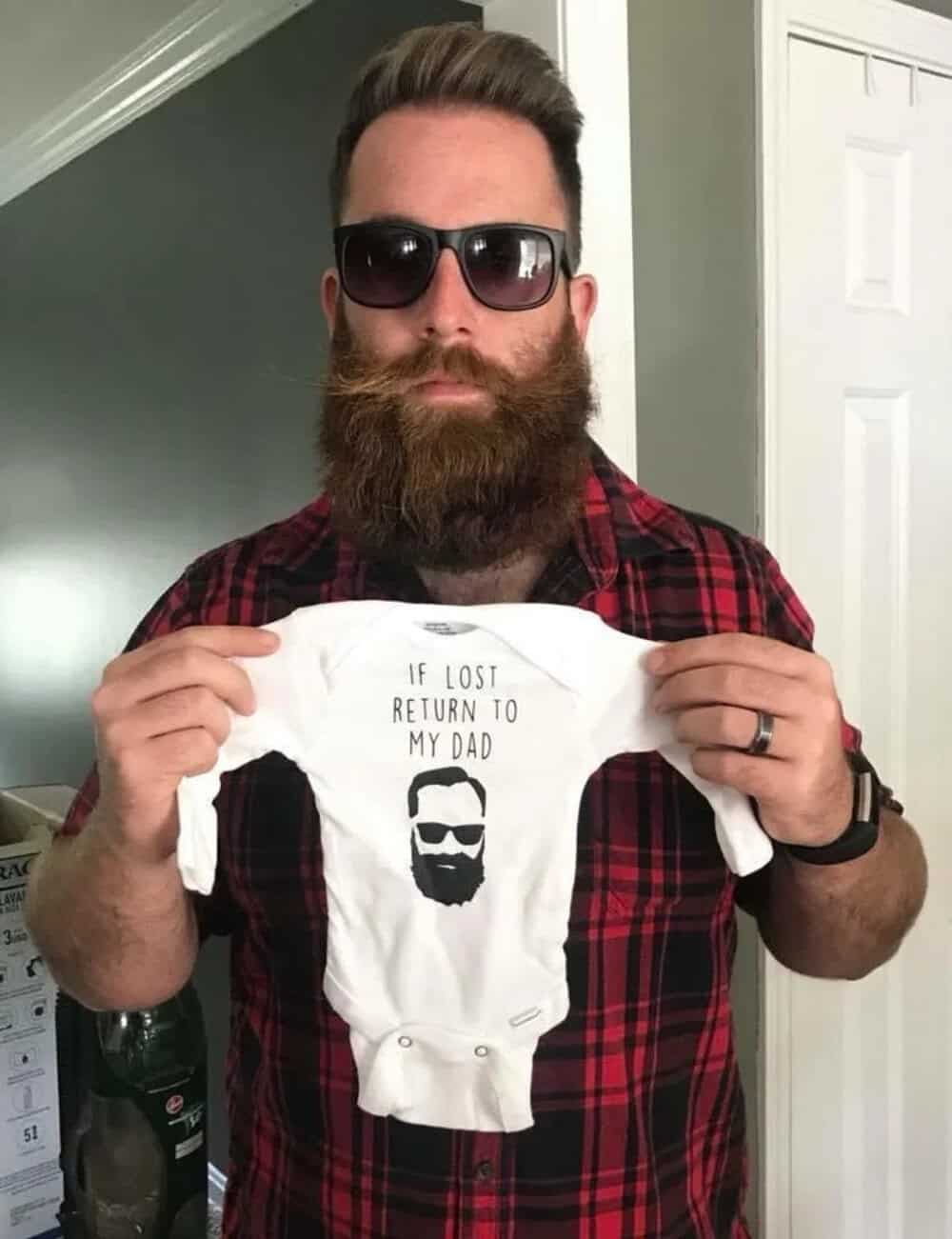 A man with a beard and sunglasses holding a baby onesie with a beard graphic and text.
