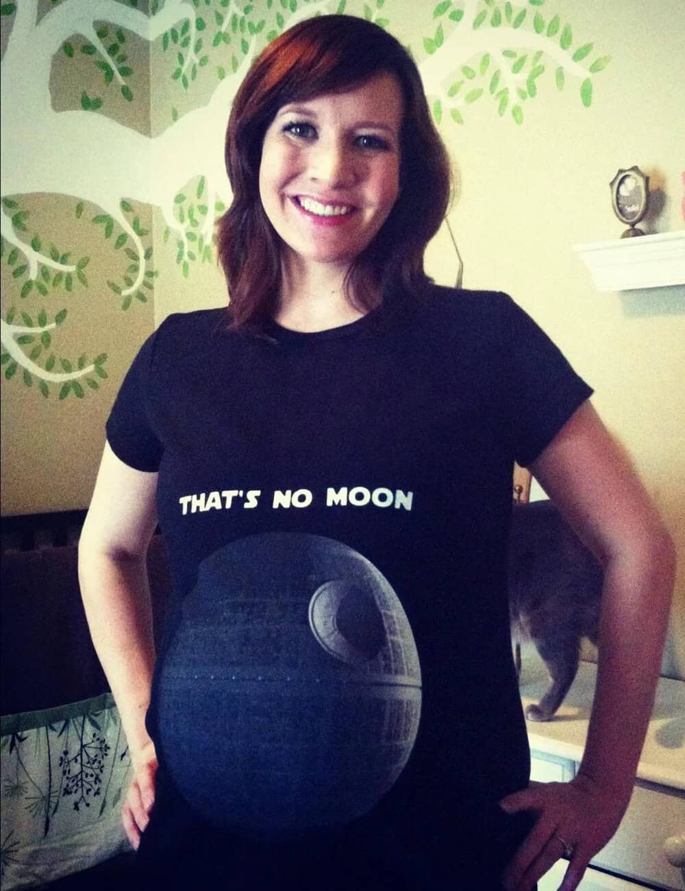 A woman with red hair smiling, wearing a black T-shirt with a moon design and the text "THAT'S NO MO.