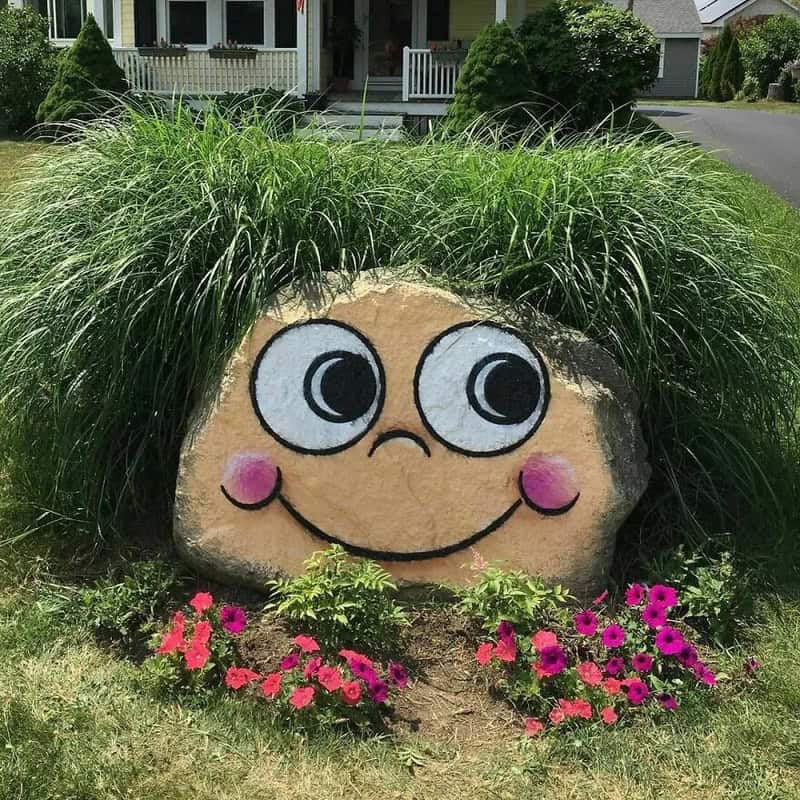 Big rock with a painted smiling face, large eyes, and pink cheeks, surrounded by pink flowers and ta.