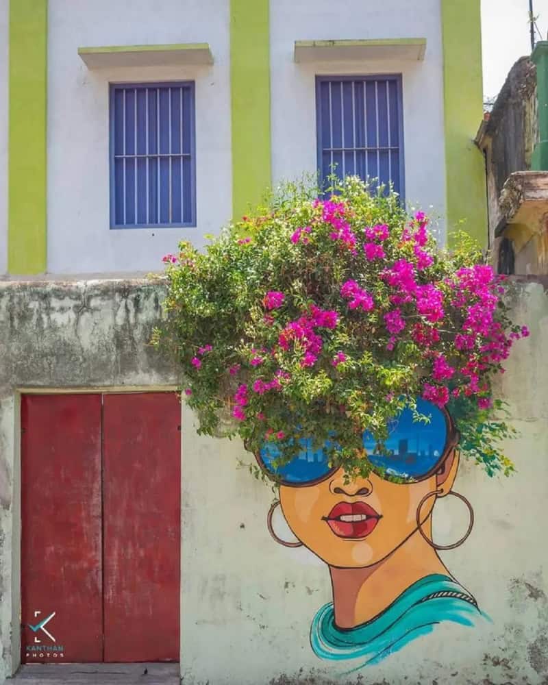 A woman with large sunglasses and hoop earrings, with a colorful flower bush as her hair, painted on.