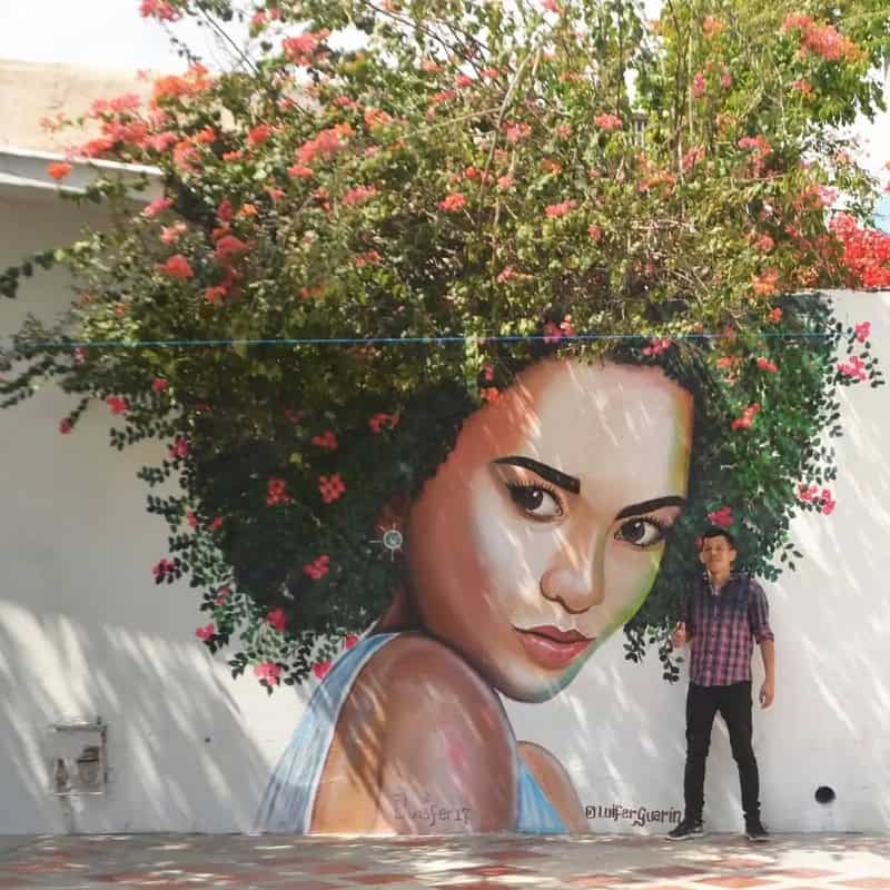 Large mural of a woman's face with floral hair on a white wall, with a man standing nearby.