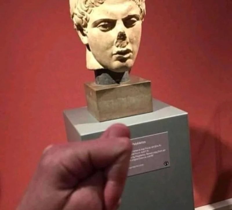 Ancient Greek sculpture bust with a hand holding a tiny object in front.
