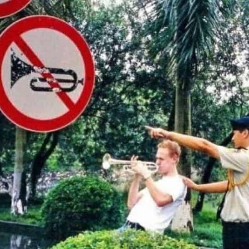No horn playing allowed sign with two boys, one playing trumpet and the other pointing, in a park se.
