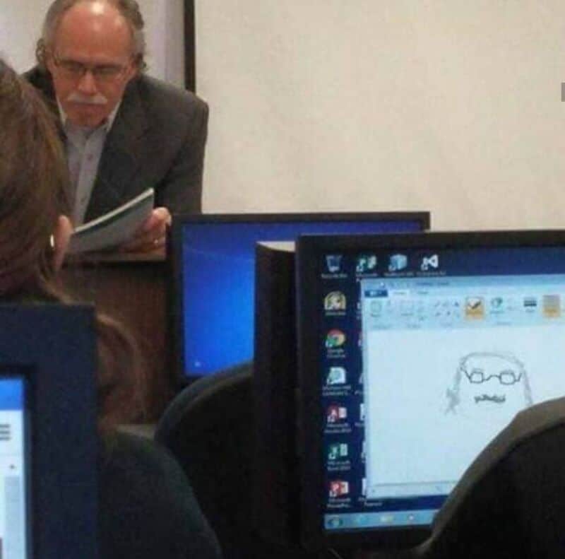 Man reading a book in front of a computer screen with a drawing of a face and glasses.