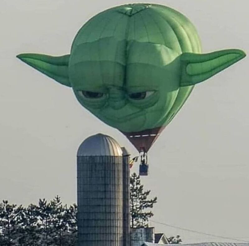 Yoda-shaped hot air balloon flying near a silo with trees in the background.