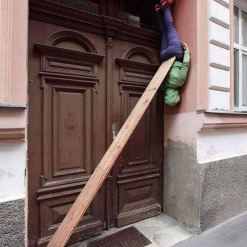 Child hanging upside down from a wooden plank against a building door.