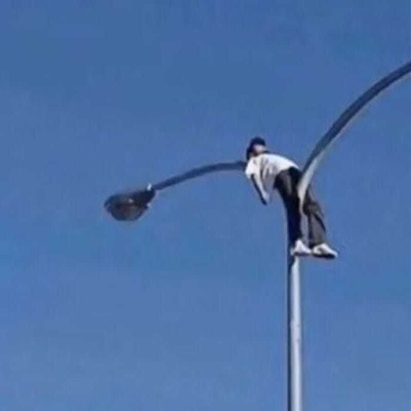 Child balancing on a streetlamp arm against a blue sky.