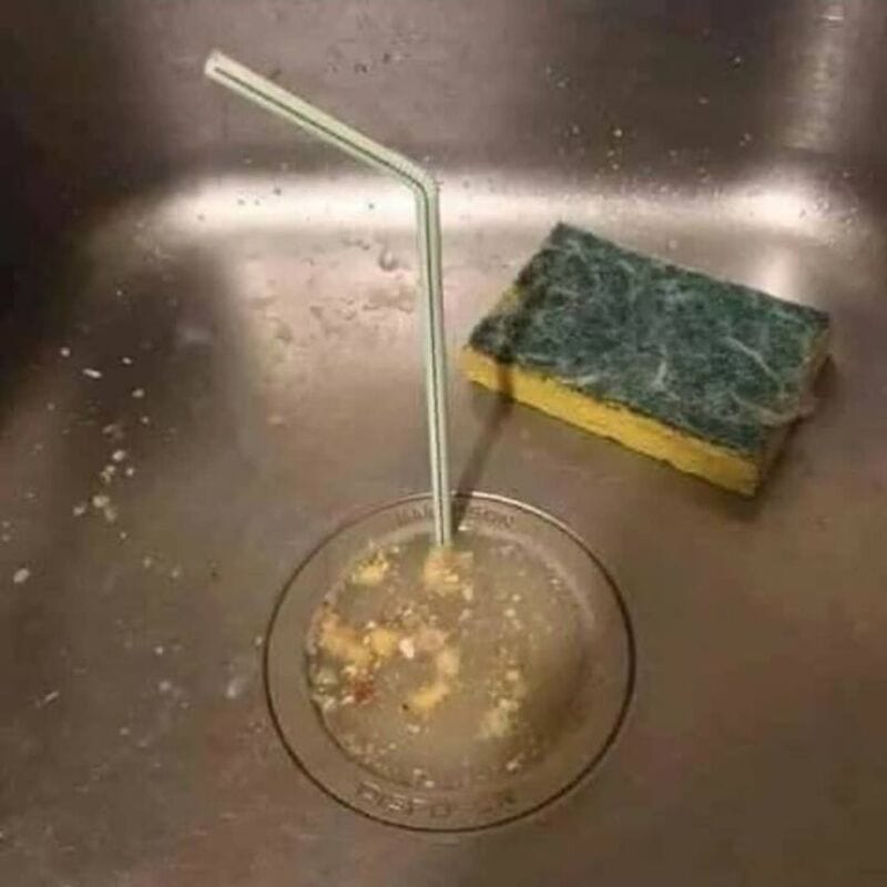 An empty glass with a straw and a cleaning sponge on a sink drain.