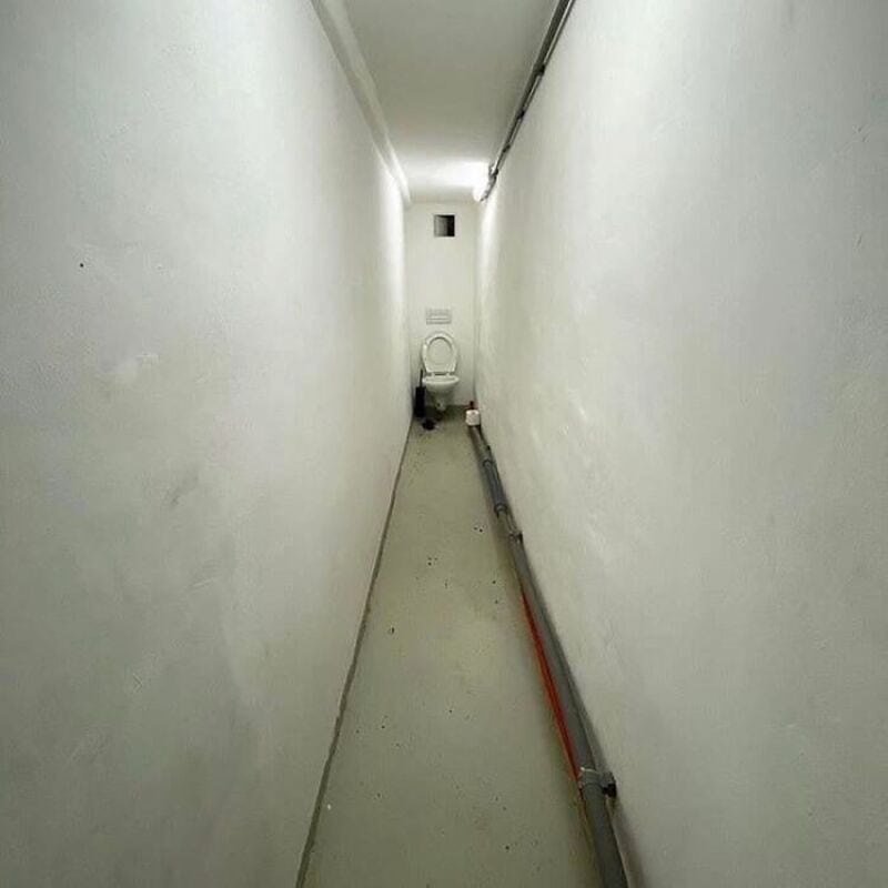 Narrow corridor with a toilet at the end, plain white walls, and a pipe along the floor.