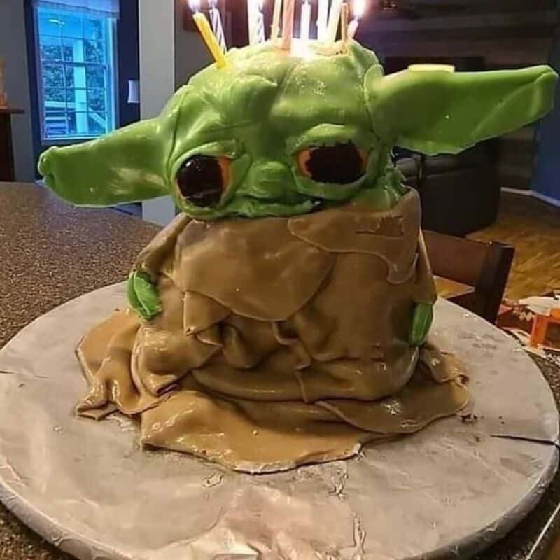 Baby Yoda cake with candles, green ears, and brown robe on a cake board.