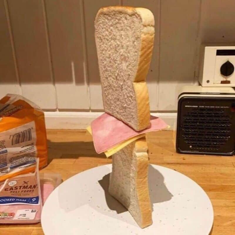 An artistic sandwich sculpture with bread slices, ham, and cheese stacked vertically on a white plat.
