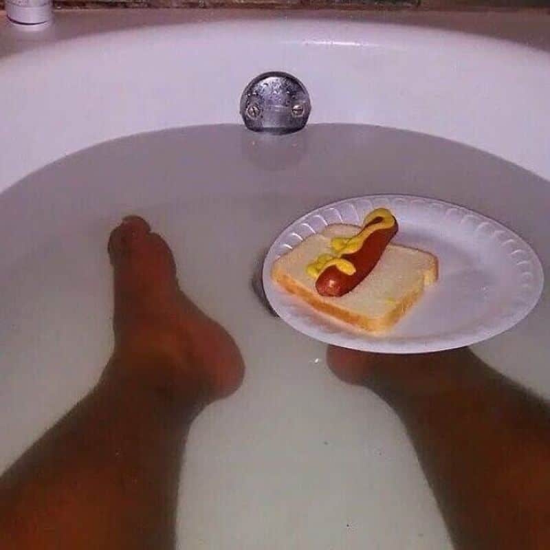 A1: Person's legs in a bathtub with a paper plate holding a hot dog and mustard.