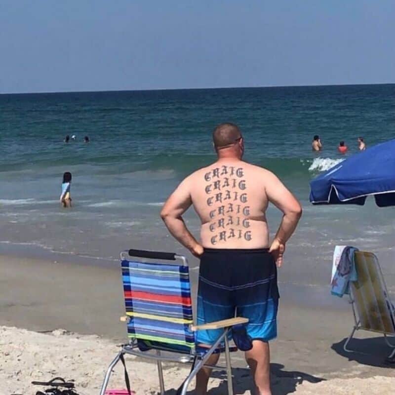 Craig tattoo on his back, standing on the beach with people swimming in the ocean.