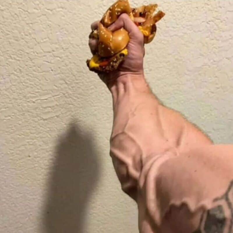 Hand gripping a large, messy cheeseburger against a beige wall.