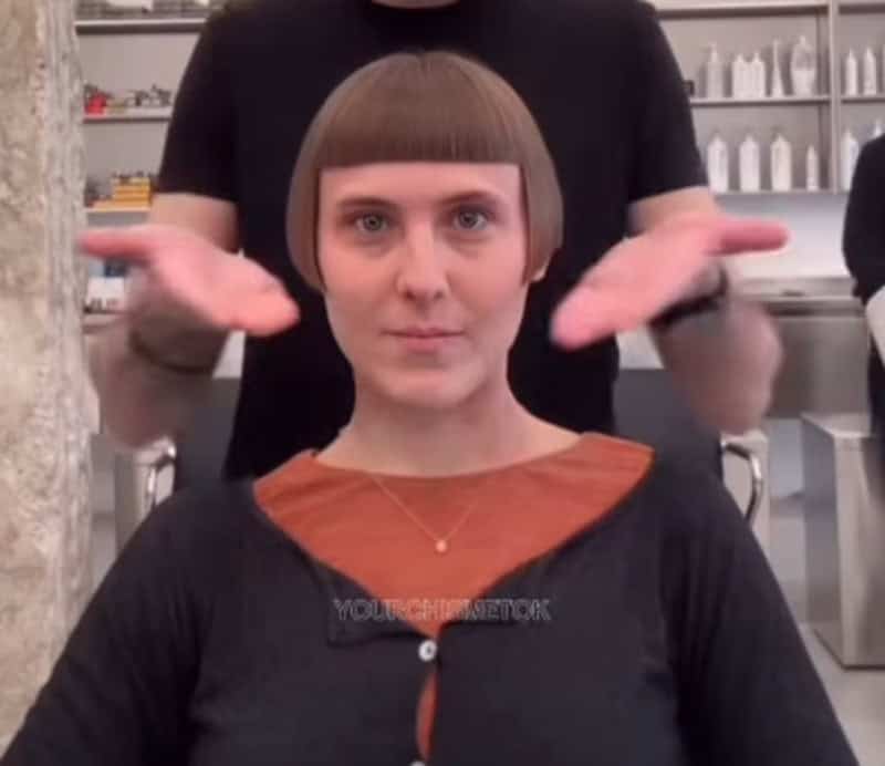Woman with a blunt, straight-across haircut in a salon, with a stylist behind her.