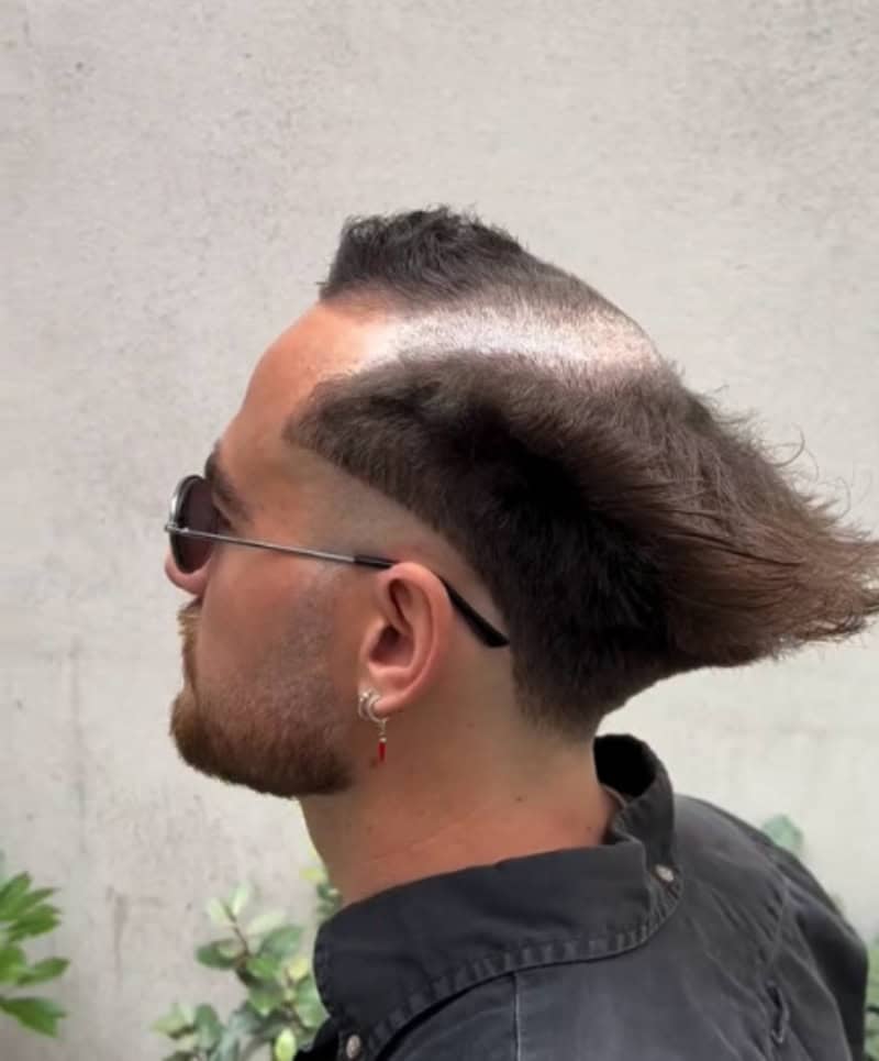 Unique hairstyle with shaved sides and long, flowing top, in front of a plain wall.