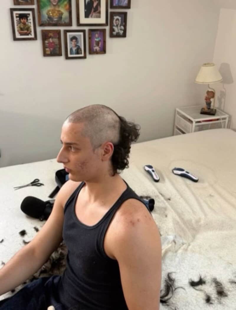 Young man with a poorly cut mohawk hairstyle, sitting on a bed.