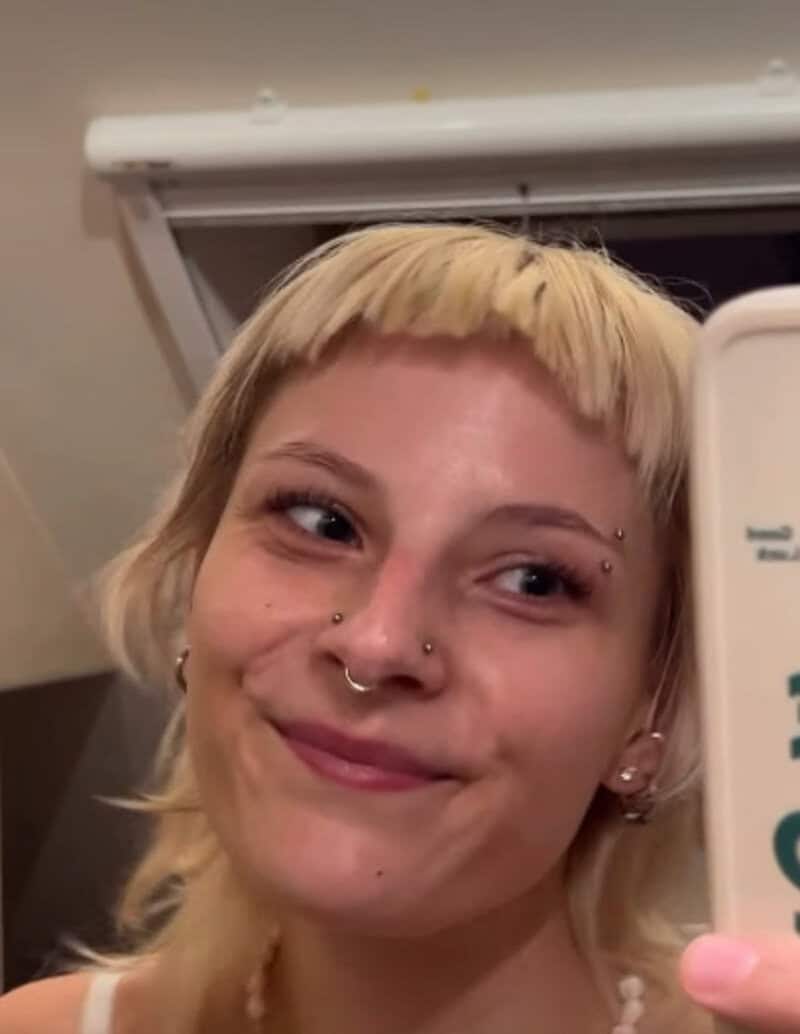 Young woman with short blonde hair, multiple facial piercings, smiling in front of a mirror.
