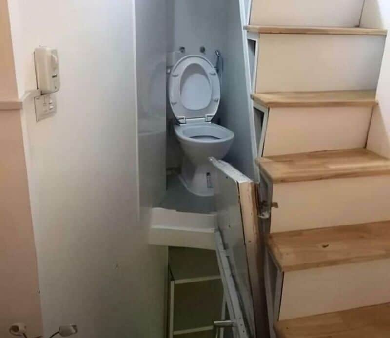 Toilet installed under staircase with a makeshift platform and partial wall.