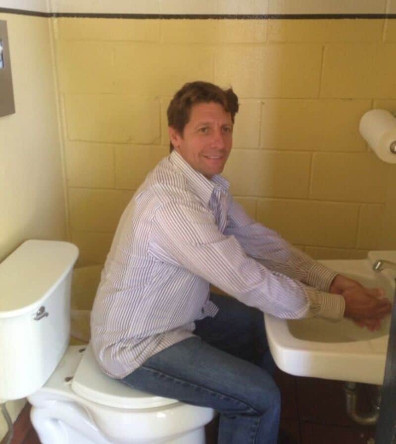 Man sitting on toilet, washing hands at sink in bathroom.